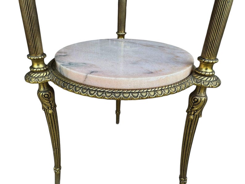 May include: An antique side table with a round, pink marble top. The table has a brass-coloured metal frame with ornate detailing and three curved legs. The table is designed to hold decorative items.