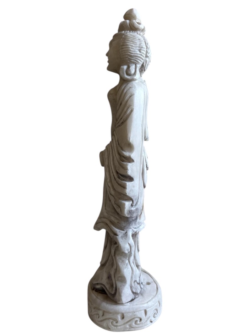 K&ouml;nnte beinhalten: Eine wei&szlig;e Statue einer Figur in flie&szlig;enden Gew&auml;ndern, die auf einem runden Sockel mit einem dekorativen Muster steht. Die Statue ist mit komplizierten Details geschnitzt, die den ruhigen Ausdruck und die elegante Kleidung der Figur zeigen. Die Statue ist ein dekorativer Gegenstand.
