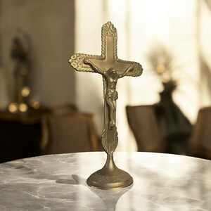May include: A gold-colored crucifix with a detailed depiction of Jesus Christ. The cross has ornate detailing and stands on a decorative base. The religious item is displayed on a marble-like surface.