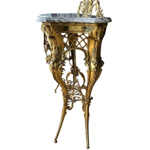 May include: Ornate gold-colored side table with a marble top. The table features intricate floral and scrollwork details, with a decorative back and slender, curved legs. The table's height is approximately 30 inches.