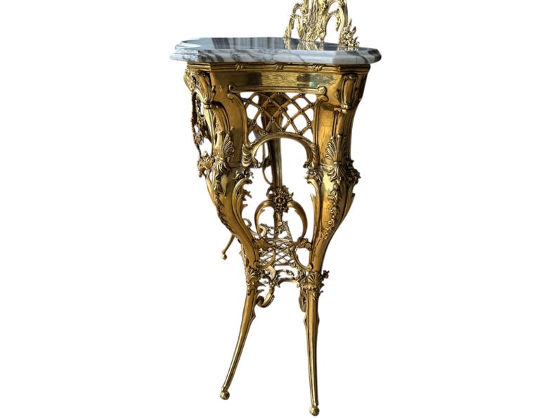 May include: Ornate gold-colored side table with a marble top. The table features intricate floral and scrollwork details, with a decorative back and slender, curved legs. The table's height is approximately 30 inches.