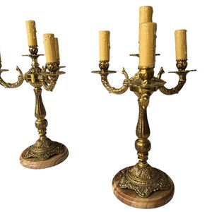 Pu&ograve; includere: Due candelabri antichi in ottone, ciascuno con cinque portacandele. I candelabri presentano dettagli ornati e poggiano su basi di marmo. Le candele sono di colore beige chiaro.