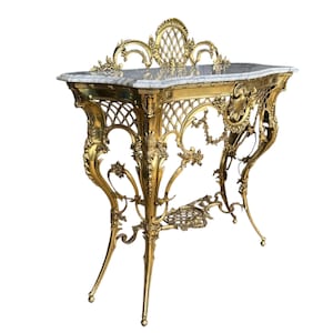 May include: An ornate, gold-colored console table with a marble top. The table features intricate, scrolling metalwork, including a lattice design on the sides and floral accents. The table's dimensions are approximately 48 inches wide, 18 inches deep, and 36 inches high.