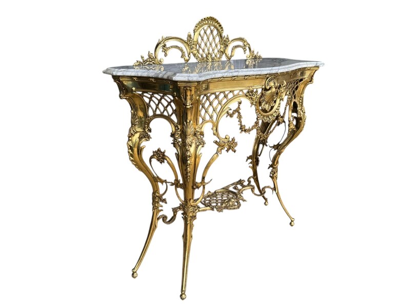 May include: An ornate, gold-colored console table with a marble top. The table features intricate, scrolling metalwork, including a lattice design on the sides and floral accents. The table's dimensions are approximately 48 inches wide, 18 inches deep, and 36 inches high.