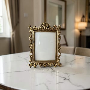 May include: A rectangular picture frame with a metallic gold finish and an ornate, raised scrollwork border. The frame features a beaded inner edge surrounding a blank white center panel, standing upright with a detailed, symmetrical design.