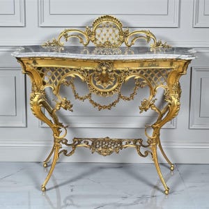 May include: A gold-colored console table with ornate detailing. The table features a marble top and intricate scrollwork on the legs and frame. The design includes a decorative back and a lower shelf.
