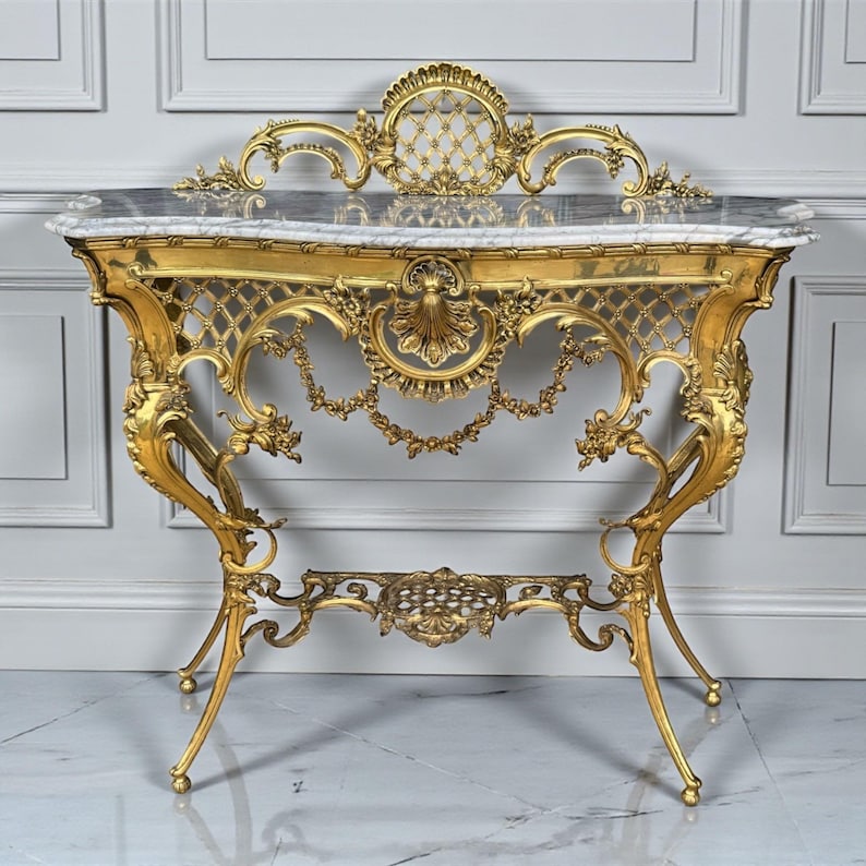 May include: A gold-colored console table with ornate detailing. The table features a marble top and intricate scrollwork on the legs and frame. The design includes a decorative back and a lower shelf.