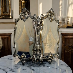 Könnte beinhalten: Verzierte, silberfarbene Wandleuchte mit zwei satinierten Glasscheiben. Die Paneele weisen ein Goldblatt-Design auf. Die Leuchte hat ein aufwendiges, dekoratives Design mit Schnörkeldetails und einem zentralen Träger.