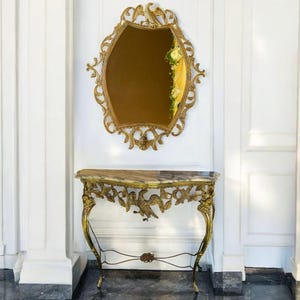 Könnte beinhalten: Verzierter goldfarbener Spiegel mit dekorativem Rahmen, über einem passenden Konsolentisch. Der Tisch hat eine Marmorplatte und aufwendige goldfarbene Metallarbeiten mit Vogel- und Blumenmotiven. Spiegel und Tisch erzeugen eine luxuriöse, antike Ästhetik.