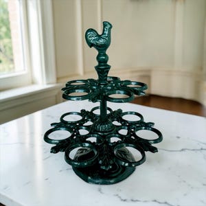 May include: A dark green cast iron egg holder, topped with a rooster figurine. The two-tiered holder features circular openings for eggs and is displayed on a white marble surface. The decorative kitchenware item is designed to hold a dozen eggs.