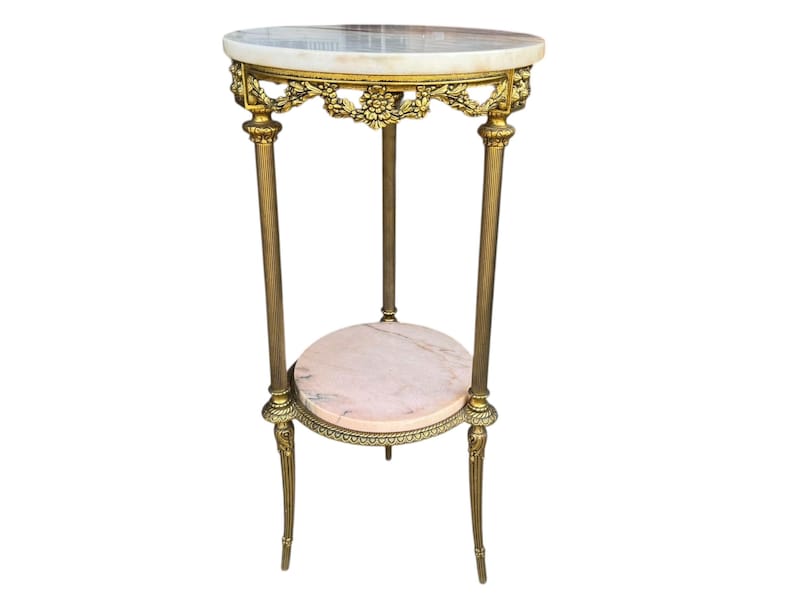 May include: An antique, two-tiered side table with a marble top and a lower shelf. The table has a gold-coloured metal frame with ornate floral detailing and slender legs. The top is approximately 61 cm in diameter.