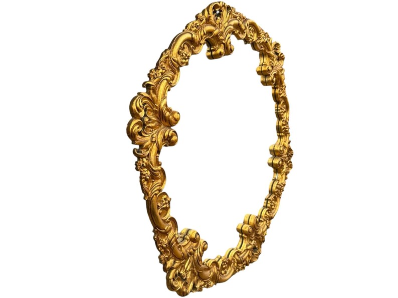 May include: Ornate gold-colored oval frame with detailed scrollwork and floral accents. The frame has a three-dimensional design and is set against a white background. The frame is likely for a mirror or artwork.