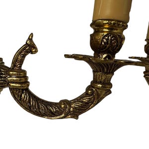 Pu&ograve; includere: Primo piano di un'applique in ottone vintage con tre portacandele. L'applique presenta bracci curvi e ornati con dettagli a foglie e volute. I portacandele sono sormontati da finte candele color crema.
