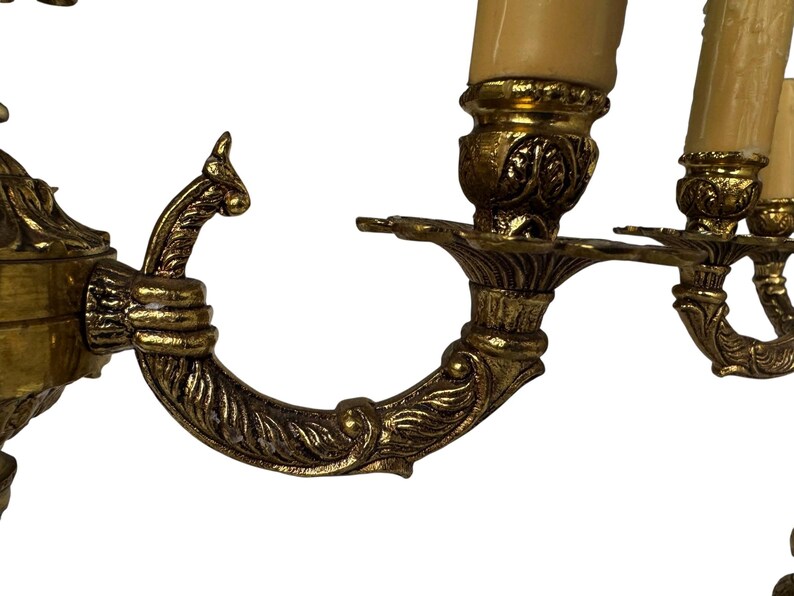 Pu&ograve; includere: Primo piano di un'applique in ottone vintage con tre portacandele. L'applique presenta bracci curvi e ornati con dettagli a foglie e volute. I portacandele sono sormontati da finte candele color crema.