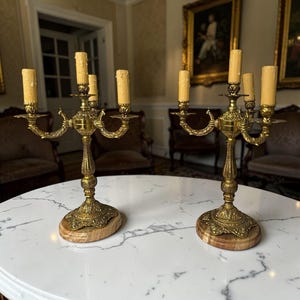 Pu&ograve; includere: Due candelabri antichi in ottone, ciascuno con cinque portacandele. I candelabri presentano disegni ornati e dettagliati e sono posti su basi ovali in marmo. Le candele sono di colore giallo crema. Su un tavolo di marmo bianco.
