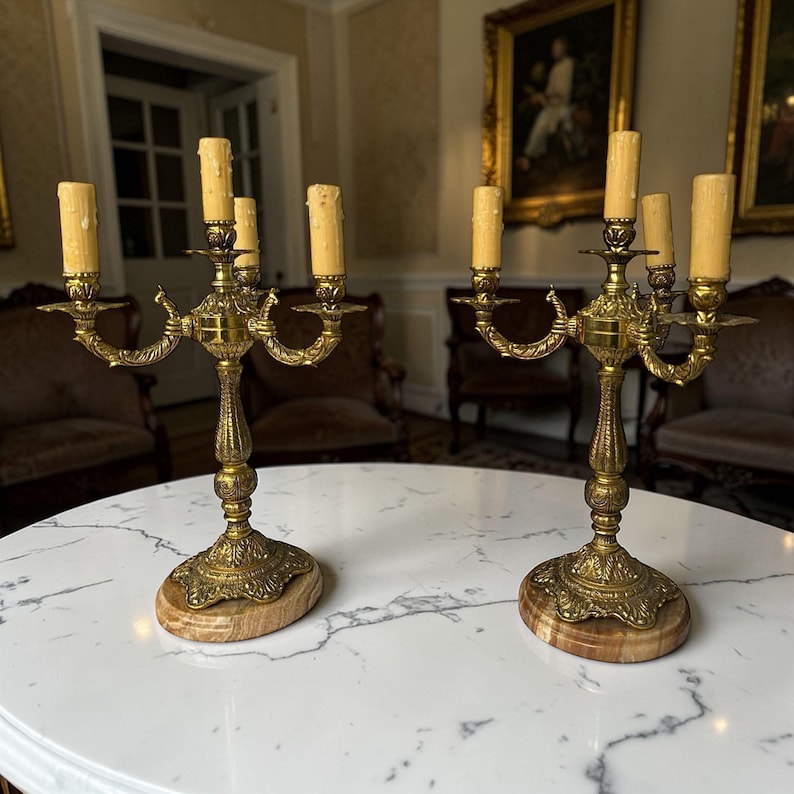 Pu&ograve; includere: Due candelabri antichi in ottone, ciascuno con cinque portacandele. I candelabri presentano disegni ornati e dettagliati e sono posti su basi ovali in marmo. Le candele sono di colore giallo crema. Su un tavolo di marmo bianco.
