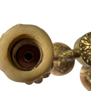 Pu&ograve; includere: Primo piano di un lampadario vintage in ottone con intricati dettagli floreali e a volute. L'apparecchio di illuminazione ha un portacandele centrale con resti di cera e un braccio decorativo. L'ottone ha una calda patina invecchiata.