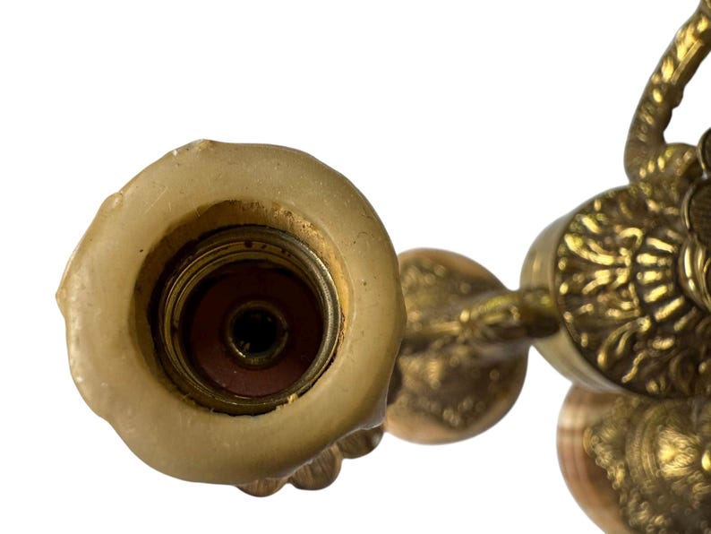 Pu&ograve; includere: Primo piano di un lampadario vintage in ottone con intricati dettagli floreali e a volute. L'apparecchio di illuminazione ha un portacandele centrale con resti di cera e un braccio decorativo. L'ottone ha una calda patina invecchiata.