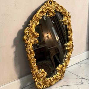 May include: Ornate, gold-colored mirror with an oval shape. The frame features intricate, swirling details and floral accents. The mirror is leaning against a neutral-colored wall and is positioned on a marble floor.