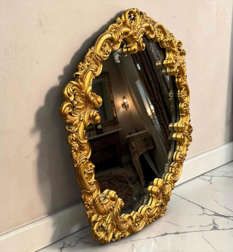May include: Ornate, gold-colored mirror with an oval shape. The frame features intricate, swirling details and floral accents. The mirror is leaning against a neutral-colored wall and is positioned on a marble floor.
