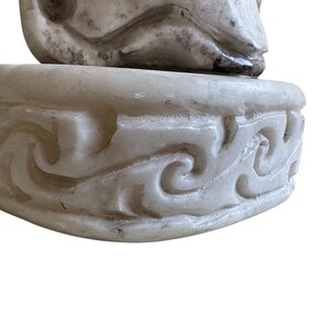 K&ouml;nnte beinhalten: Eine geschnitzte, cremefarbene Skulptur mit detailliertem Sockel. Der Sockel weist ein sich wiederholendes, wirbelndes Muster auf. Die Skulptur scheint aus Stein oder einem &auml;hnlichen Material zu bestehen, mit sichtbarer Textur und leichter Verf&auml;rbung.