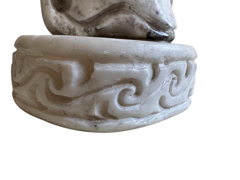 K&ouml;nnte beinhalten: Eine geschnitzte, cremefarbene Skulptur mit detailliertem Sockel. Der Sockel weist ein sich wiederholendes, wirbelndes Muster auf. Die Skulptur scheint aus Stein oder einem &auml;hnlichen Material zu bestehen, mit sichtbarer Textur und leichter Verf&auml;rbung.