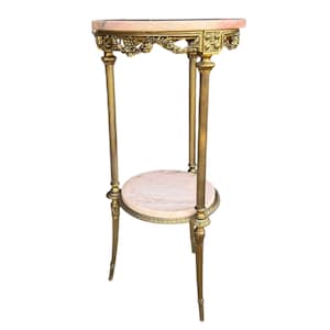 May include: An antique gold-toned plant stand with two marble shelves. The top shelf is oval-shaped and the bottom shelf is round. The stand has ornate detailing and is approximately 76 cm tall.