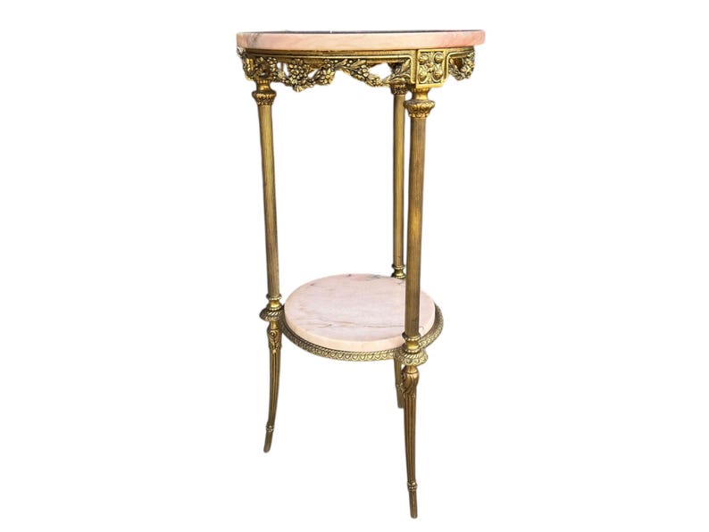 May include: An antique gold-toned plant stand with two marble shelves. The top shelf is oval-shaped and the bottom shelf is round. The stand has ornate detailing and is approximately 76 cm tall.