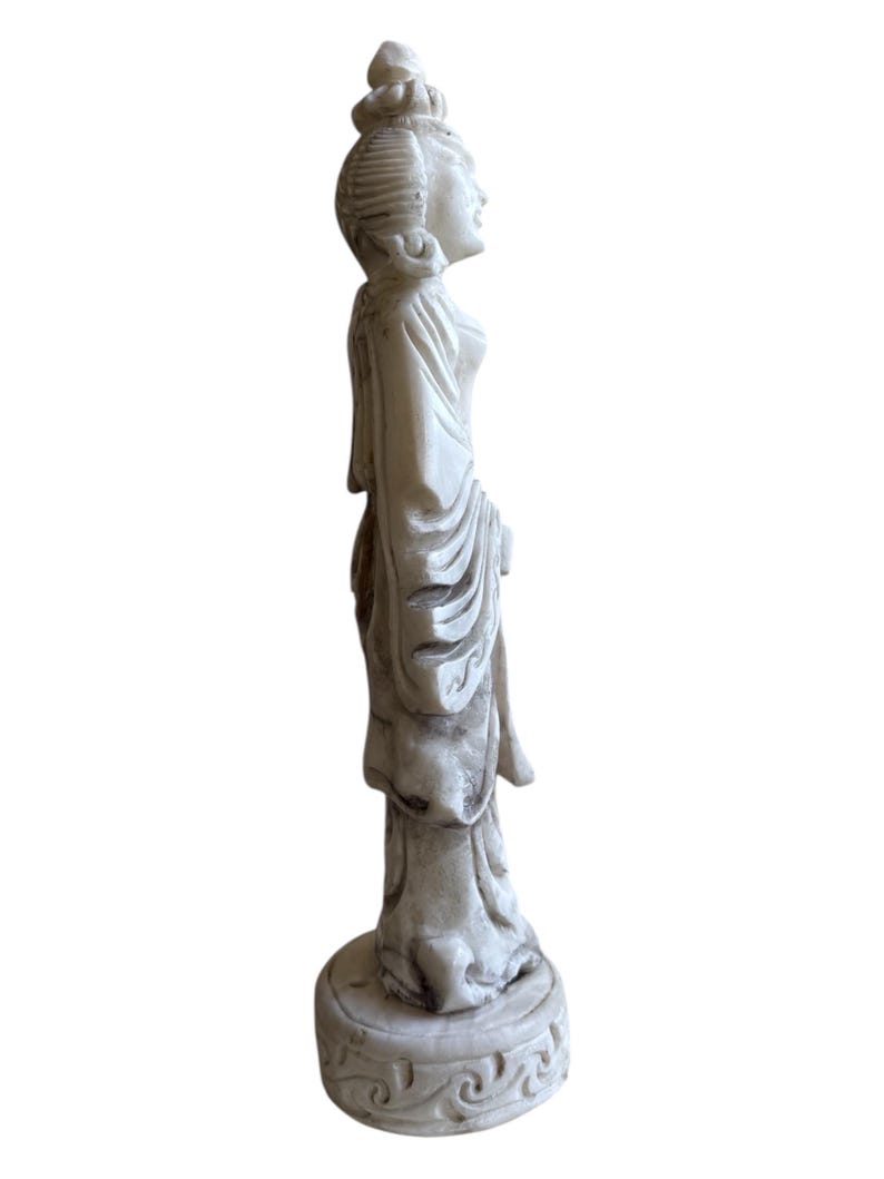 K&ouml;nnte beinhalten: Cremefarbene Statue einer Frau in flie&szlig;endem Gewand, stehend auf einem runden Sockel mit dekorativem Muster. Die Statue hat detaillierte Gesichtsz&uuml;ge und eine traditionelle Frisur. Die Figur ist im Seitenprofil, das das elegante Design zeigt.
