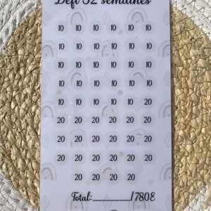 Peut inclure: Une carte blanche avec un design noir et gris. La carte porte le texte "Défi 52 semaines" et une grille de nombres, dont 10 et 20. La carte porte également le texte "Total : 1780€".