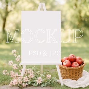 Puede incluir: Un caballete blanco con un letrero en blanco, la palabra "MOCKUP" está escrita en el letrero. Una cesta de manzanas rojas se encuentra sobre una tela blanca. Flores rosas en primer plano. El fondo es una escena exterior verde borrosa.
