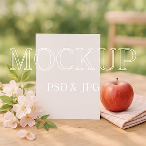 Op de afbeelding: Een blanco witte mockupkaart met het woord "MOCKUP" en "PSD & JPG" erop gedrukt. Een rode appel ligt op een roze geruite servet, met roze bloemen en groene bladeren links. De scène speelt zich af op een houten tafel.