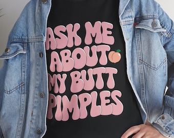 May include: A black t-shirt featuring the pink text ASK ME ABOUT MY BUTT PIMPLES and a small peach graphic, worn under a light blue denim jacket. The shirt has a crew neckline and short sleeves, with the text displayed in a bold, rounded font.