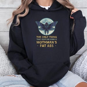 Peut inclure: Un sweat-shirt à capuche noir avec un motif de papillon de nuit centré dans une lune pâle. Sous le papillon, un texte doré indique : THE ONLY THING THAT KEEPS ME GOING IS MOTHMAN'S FAT ASS. Le sweat possède une poche kangourou et des manches longues.