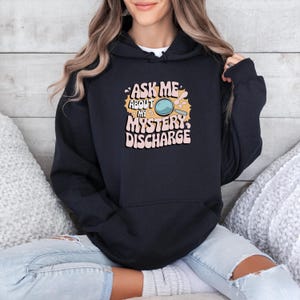Peut inclure: Sweat-shirt &agrave; capuche noir avec un imprim&eacute; graphique d'une loupe et le texte "Ask me about my mystery discharge".