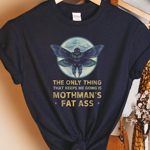 Pode incluir: Uma camiseta azul marinho com um gráfico de um mothman com olhos vermelhos brilhantes na frente de uma lua cheia. O texto "The only thing that keeps me going is Mothman's Fat Ass" é impresso em dourado abaixo do gráfico.