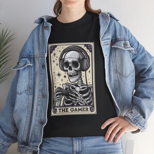 May include: Black t-shirt featuring a skeleton wearing headphones and holding a game controller. The design is framed with a decorative border and the text "THE GAMER" at the bottom. The skeleton is set against a background of stars.