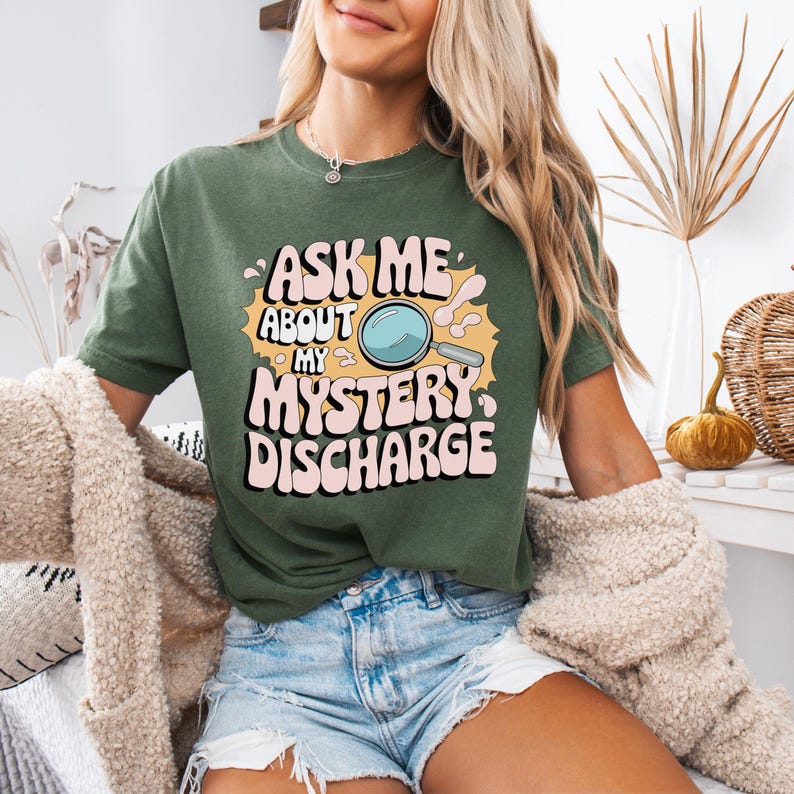 Peut inclure: Un t-shirt vert avec un graphique de style r&eacute;tro qui dit "Ask Me About My Mystery Discharge" avec une loupe et une bulle de dialogue de dessin anim&eacute;.