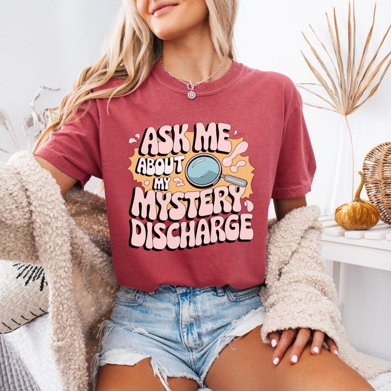 Peut inclure: Un t-shirt rouge avec le texte "Ask me about my mystery discharge" dans une police r&eacute;tro. Une loupe est &agrave; c&ocirc;t&eacute; du texte.