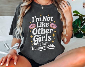 May include: Dark gray t-shirt with the text "I'm Not Like Other Girls I Have Hemorrhoids" in white, with pink donut graphics. The shirt has a vintage, distressed look.
