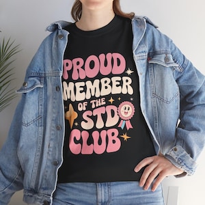 May include: Black t-shirt with pink text that reads "Proud Member of the STD Club". The text is in a retro font style. A pink cartoon character with a ribbon is above the word "Club".
