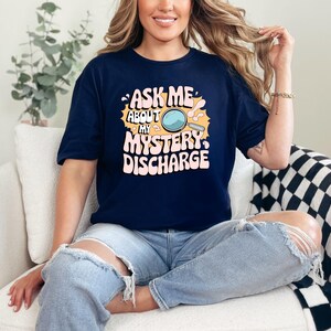 Peut inclure: Un t-shirt bleu marine avec un graphique d'une loupe et le texte "Ask Me About My Mystery Discharge" dans une police r&eacute;tro.