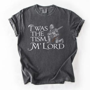 May include: A dark gray t-shirt with a distressed look features a white knight kneeling with a sword and the text "Twas the tism, M'Lord".