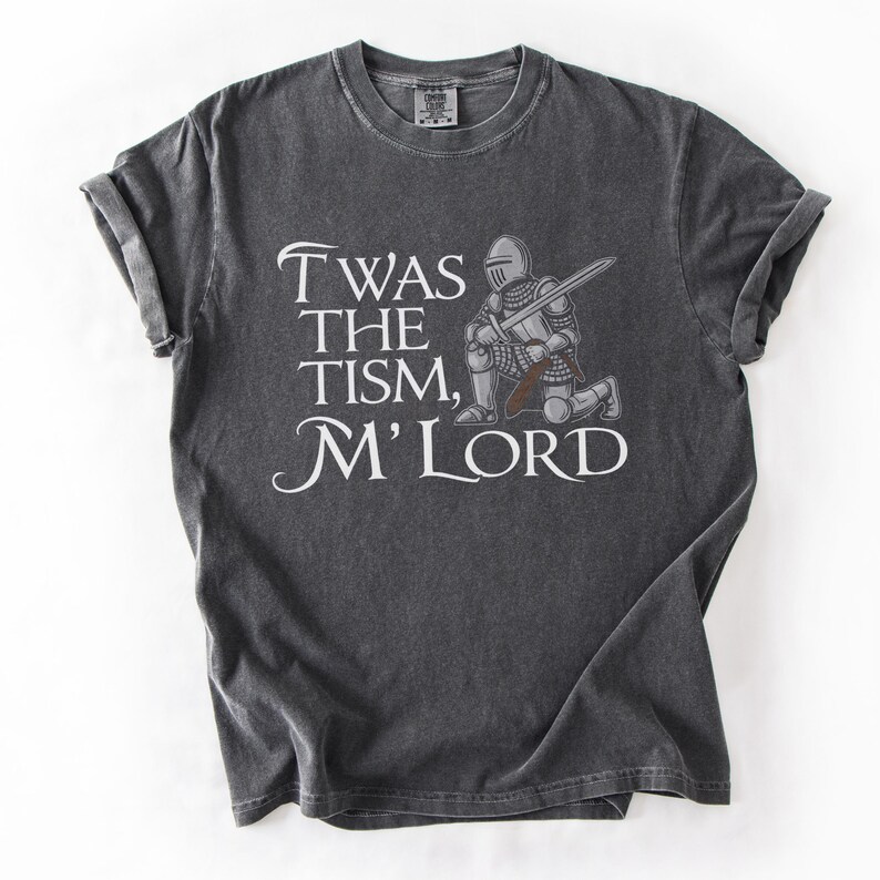 May include: A dark gray t-shirt with a distressed look features a white knight kneeling with a sword and the text "Twas the tism, M'Lord".