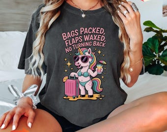 May include: Dark gray t-shirt with a graphic of a unicorn wearing sunglasses and a bikini, holding a drink and a suitcase. The text on the shirt reads "BAGS PACKED, FLAPS WAXED, NO TURNING BACK".