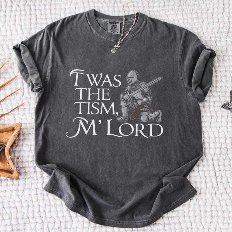 May include: A dark gray t-shirt with a distressed look features a knight kneeling with a sword and the text "Twas the tism, M'Lord".