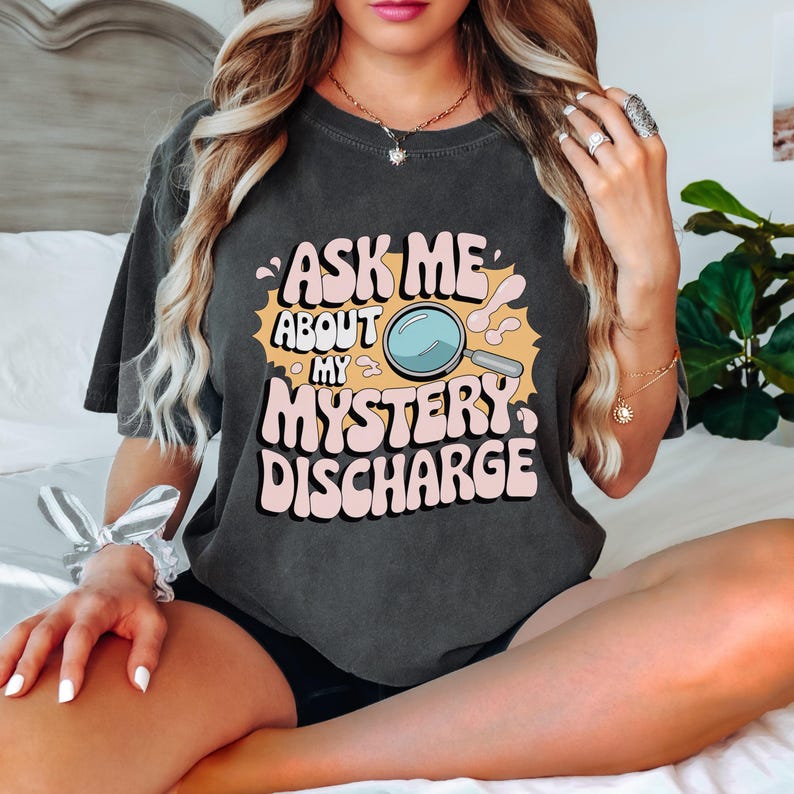 Peut inclure: Un t-shirt gris fonc&eacute; avec un imprim&eacute; graphique qui dit "Ask Me About My Mystery Discharge" dans une police r&eacute;tro. Le graphique comprend une loupe de dessin anim&eacute;.