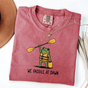 May include: A dusty rose short-sleeve t-shirt featuring a graphic of a green frog wearing a yellow life vest, holding a yellow paddle above its head and carrying a yellow dry bag. The text below the frog reads, "WE PADDLE AT DAWN."