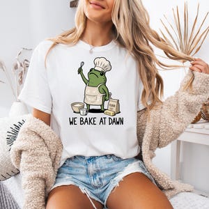 May include: A white t-shirt featuring a graphic of a green frog wearing a chef hat and apron, holding a whisk. The frog is surrounded by a bowl, a measuring cup, a bag of flour, and a stick of butter. The text "WE BAKE AT DAWN" is printed below.