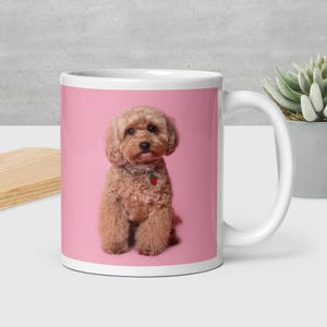 May include: White ceramic mug with a pink background featuring a small, light brown dog with curly fur. The dog wears a collar with a red strawberry charm. The mug is next to a small succulent plant in a gray pot.